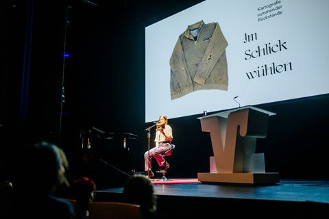 Laura Wiemers während einer Lesung im Rahmen der Preisverleihung am 21. September 2025 in Paderborn; Foto: VerbundVolksbank OWL eG Abschlussarbeit ausgezeichnet in der Kategorie »Literatur/Sprache« Laura Wiemers sitzt auf einer Bühne und hält ein Buch in der Hand, aus dem sie liest