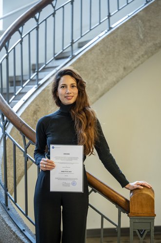 Betül Peker on the stairs in the foyer of the main building, holding her award in front of her.