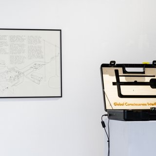 Ausstellungsansicht: Ein Koffer mit dem Text "Global Consciousness Interface" steht neben einer Zeichnung eines Geräts 