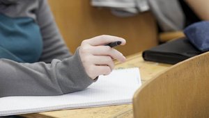 Bild im Hörsaal mit Notizblock und Hand mit Stift im Fokus