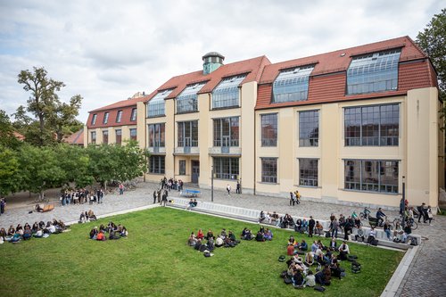 Campus mit Hauptgebäude und Studierenden, die auf der Wiese sitzen