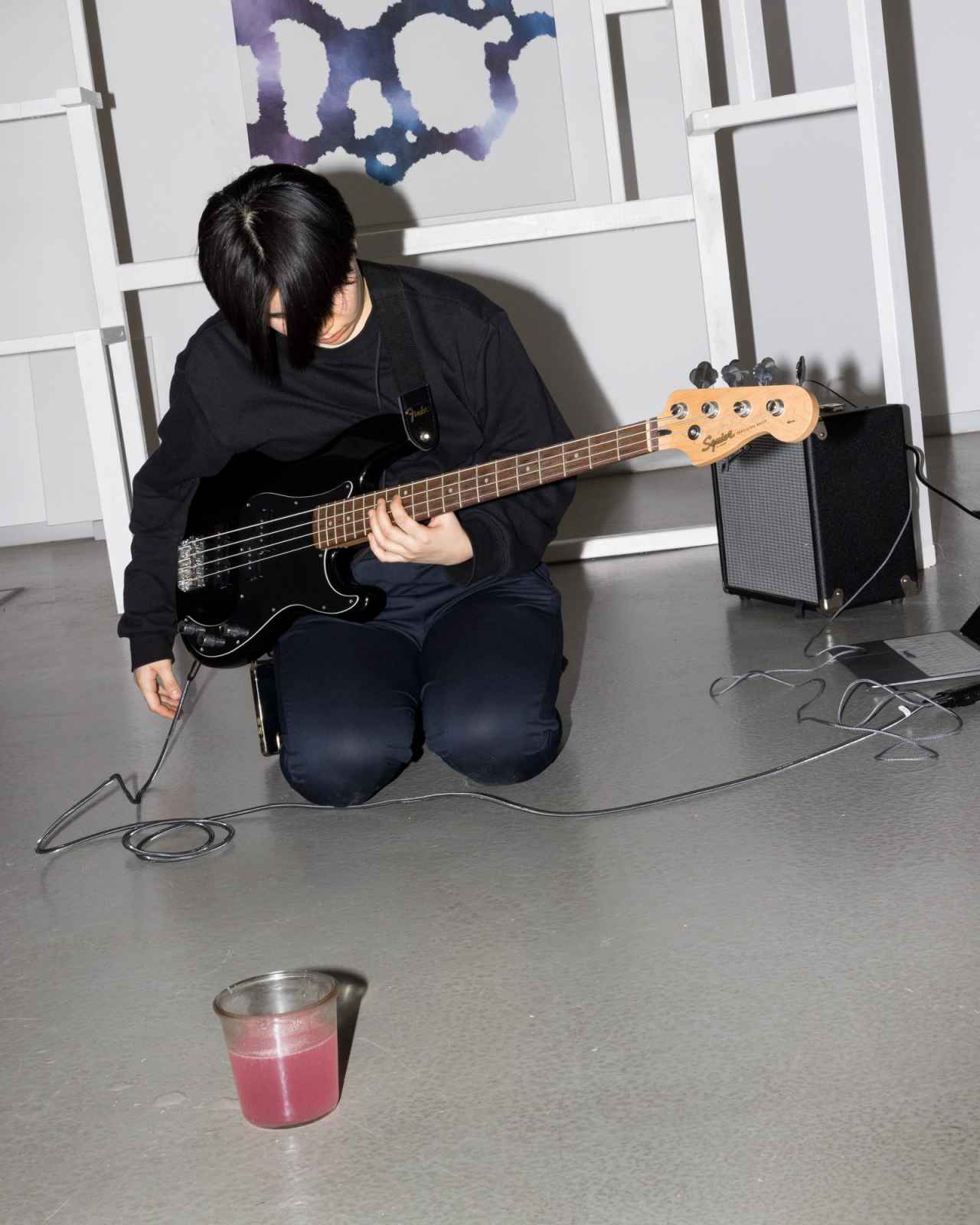an individual playing a bass guitar while kneeling on the floor and a jar filled with liquid in the foreground