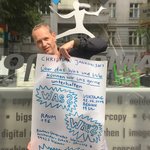 Christian Jankowski vor einem Schaufenster präsentiert ein handgeschriebenes, selbst gestaltetes Plakat zur Veranstaltung