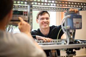 Mann steht an einer technischen Apparatur und schaut in die Kamera