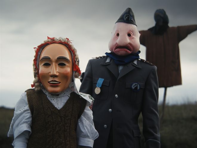 Zwei Personen im Vordergrund, im Hintergrund eine Vogelscheuche auf einem Feld.