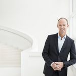 Dr. Thomas Rabe trägt einen schwarzen Anzug und steht in einem weißen hellen Raum. Er lächelt frontal in die Kamera.