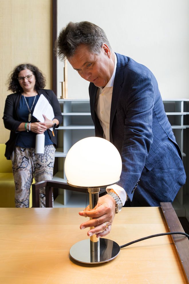 Jürgen Dusel ertastet die Wagenfeldlampe auf dem Schreibtisch im Gropiuszimmer. Im Hintergrund steht Michaela Albrecht. Er spricht lächelnd. 