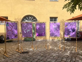 Das Bild zeigt fünft lila-farbene Wendeposter in Holzrahmen vor dem Haus der Studierenden M18.