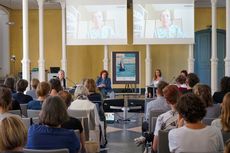 Das Bild zeigt die Aula der Uni Greifswald während einer Podiumsdiskussion. Im Hintergrund sitzen die Moderatorin und 3 Diskutantinnen unter zwei Leinwänden, die das Video-Bild der vierten Diskutantin zeigen. Im Vordergrund sind mehrere Stuhlreihen gefüllt mit Gästen.