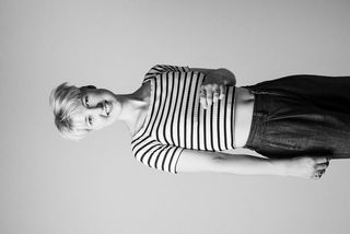 Schwarz-Weiß-Porträt von Carmen Draxler. Sie hat kurze, blonde Haare und trägt eine dunkle, weite Hose sowie ein gestreiftes T-Shirt. Sie lächelt in die Kamera.