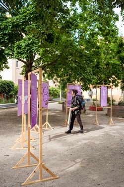 Das Foto zeigt die fünf von der Projektgruppe »Geschlechterlektüren« erstellten Wendeplakate in der Freifläche zwischen Hauptgebäude, Campus.Coffice und dem Haus der Studierenden M18. Eine Person mit kinnlangen dunklen Haaren, dunkler Kleidung und Rucksack tritt interessiert an eines der Plakate heran.