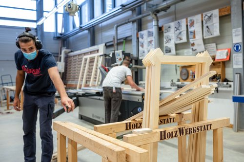 In the woodworking shop, a student wearing mouth and hearing protection points to parts for the furniture for the cafeteria foyer that will soon be assembled. Another student can be seen in the background. Bauhaus-Universität Weimar, Photo: Thomas Müller