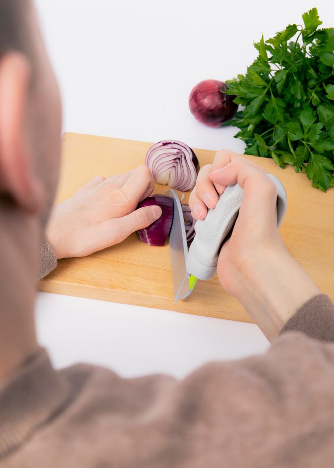 Eine Person schneidet eine rote Zwiebel auf einem Holzbrett mit einem Messer, das mit einem ergonomisch geformten, individualisierbaren Griff ausgestattet ist; im Hintergrund liegen weitere Zwiebeln und Petersilie.