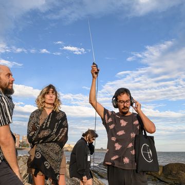 Vier Menschen stehen am Meer, einer hält ein tragbares Radio mit ausgefahrener Antenne in die Höhe und lauscht über Kopfhöhrer, die er trägt