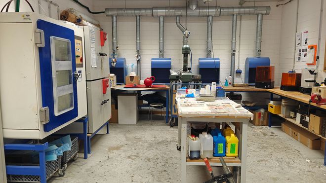 Werkraum mit mehreren Arbeitsplätzen, Absauganlagen und Materialbehältern. In der Mitte steht ein Arbeitstisch mit Werkzeugen und Behältern.