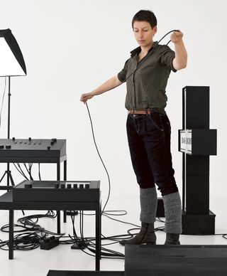 Portrait of Prof. Jana Gunstheimer. She holds a long black cable in her hands while standing among minimalist black objects and cables that are part of an installation in a bright studio.