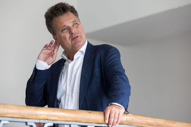 Jürgen Dusel in Nahaufnahme am Teppengeländer im Hauptgebäude: Er sieht sehr interessiert aus, hält sich eine Hand ans Ohr, als Geste, die zeigt, was er hört, und scheint zu sprechen.