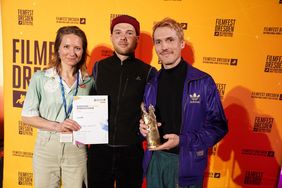 Zwei Männer und eine Frau stehen vor einer gelben Fotowand beim Filmfest Dresden. Die Frau hält eine Urkunde, einer der Männer eine Trophäe.