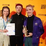 Two men and one woman stand in front of a yellow backdrop at the Dresden Film Festival. The woman holds a certificate, one of the men a trophy.