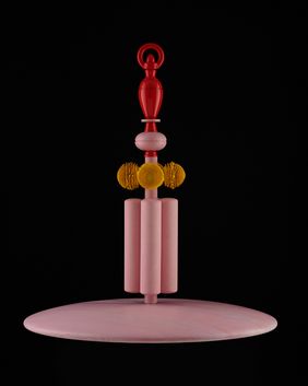 Sculptural object composed of stacked wooden elements in pink, red, and yellow tones, standing upright on a circular base. The object is centered against a black background and lit from above.