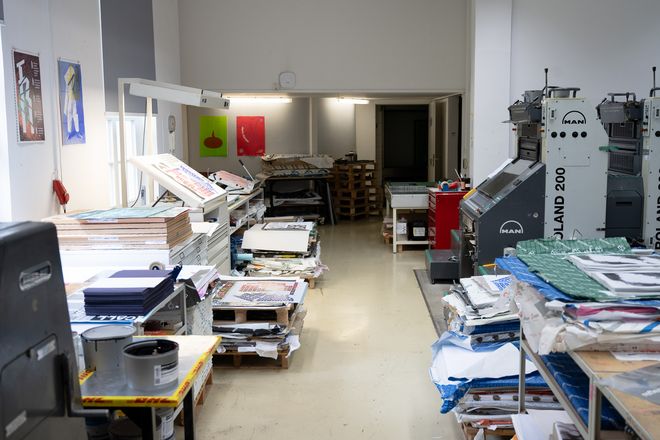 Blick in die Druckwerkstatt mit großen Druckmaschinen, Papierstapeln und Arbeitstischen. Im Vordergrund liegen Druckbögen, im Hintergrund stehen Paletten mit Papier.
