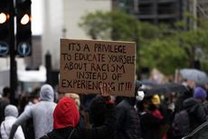 The photo shows a crowd of demonstrators. In the foreground, a Black person, shown only from behind, is holding up a sign which reads: "It's a priviledge to educate yourself about racism instead of experiencing it." The person - probably a woman - is wearing a black jacket, red nailpolish and a red sweater-hood covering their head.