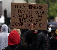 The picture shows an anti-racist protest. One of the protesters holds up a cardboard-sign with the message: It's a privilege to educate yourself about racism instead of experiencing it!!!