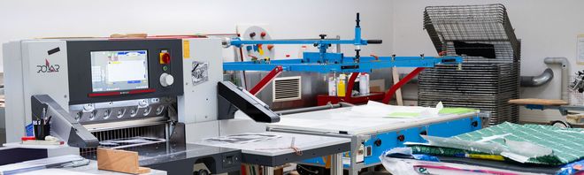 Panoramaaufnahme der Druckwerkstatt mit Schneidemaschine, Siebdruckanlage und Trockenregalen im Überblick.