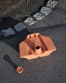 Terracotta-colored molded object lying on dark asphalt next to a curb made of granite stones. The object has a geometric shape with a central circular opening; a small matching terracotta piece lies nearby.