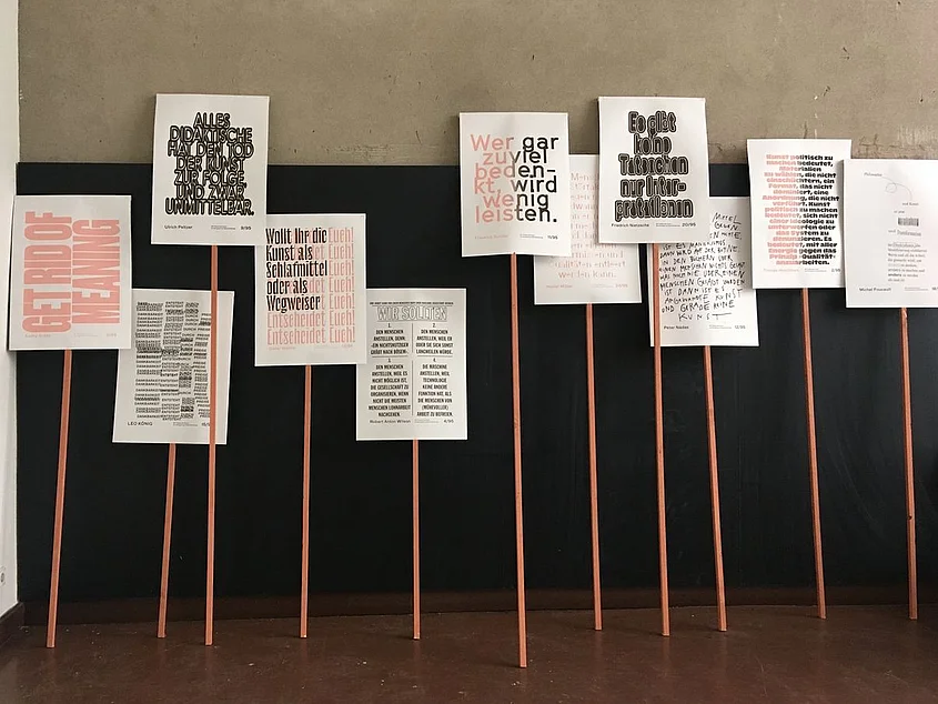 Ausstellungsansicht der Arbeit »95 Thesen der Kunst« mit typografisch gestalteten Texttafeln auf schmalen Holzstäben vor einer schwarzen Wand.