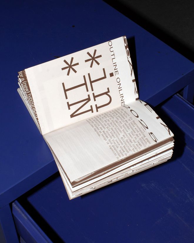 Open publication on a blue surface. The left page shows large asterisks and the letter sequence »*in«; the right page contains text with typographic markings.