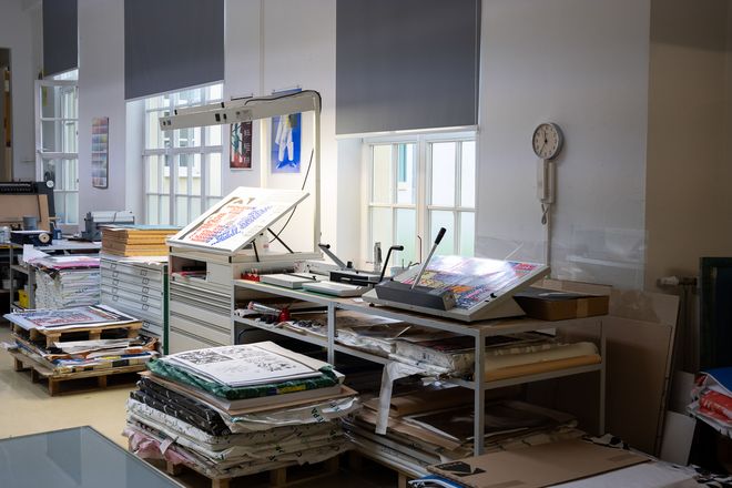 Arbeitsplatz mit Belichtungseinheit und aufgelegten Druckbögen; daneben gestapelte Papierreserven und Materialien.