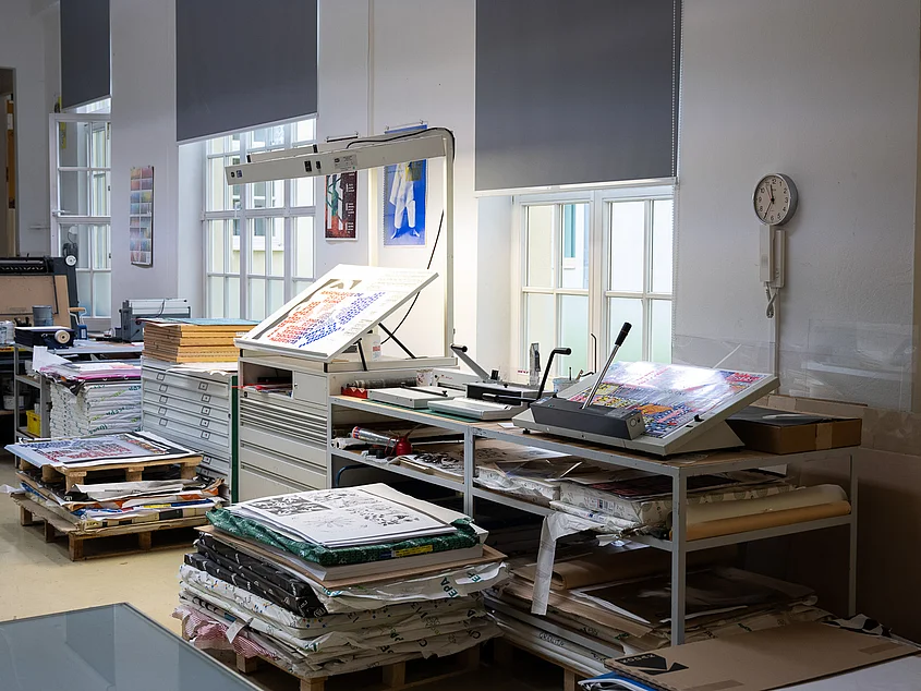 Arbeitsplatz mit Belichtungseinheit und aufgelegten Druckbögen; daneben gestapelte Papierreserven und Materialien.