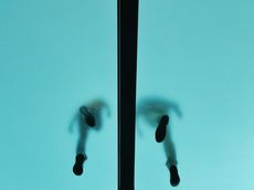 The photo shows a bluish glass ceiling from below. Through the glass, one can see the black-and-grey silhouettes of two people, probably men, standing on the glass floor.