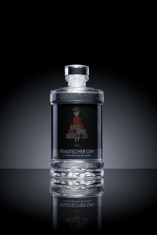 A gin bottle stands against a dark background on a reflective surface. The label shows an abstract, colourful figure made of vertical lines.