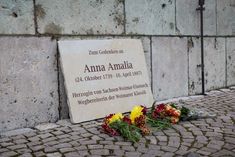 Larissa Barths Gedenktafel für Anna Amalia lehnt an der Außenmauer der Herzogin-Anna-Amalia-Bibliothek. Auf dem Kopfsteinpflaster davor liegen rote und gelbe Blumen. Die Inschrift der Kalksandstein-Tafel lautet: Zum Gedenken an Anna Amalia (24. Oktober 1739 - 10. April 1807) / Herzogin von Sachsen-Weimar-Eisenach / Wegbereiterin der Weimarer Klassik.