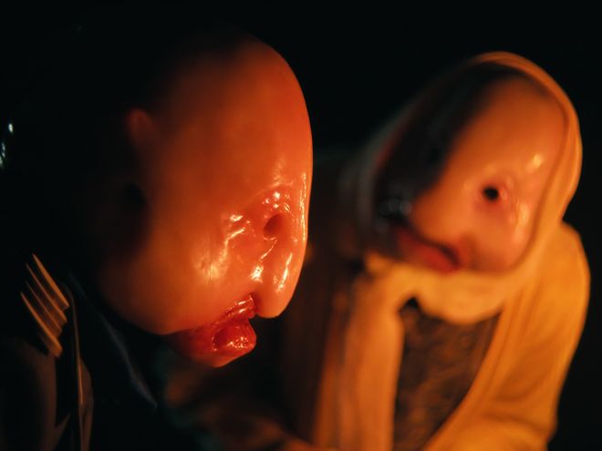 Zwei Personen mit glatten, puppenartigen Gesichtsformen im warmen Licht, eine im Vordergrund, eine leicht unscharf dahinter.