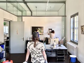 Zwei Personen mit weißen Baumwollhandschuhen richten zwei gerahmte Fotografien an einer Wand aus, eine weitere Person mit Abstand gibt Auskunft, ob die Bilder gerade hängen