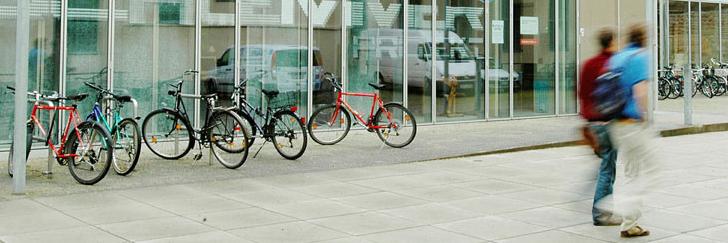 Picture of a glas building with bicycles in front of it. Two students are passing the building.