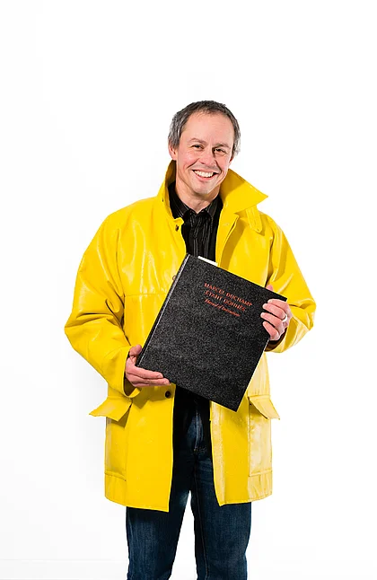 Michael Lüthy in a yellow coat against a white background. He is holding the book »Marcel Duchamp: Étant donnés« in his hands.
