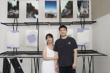 Two people standing in front of works of art and looking into the camera