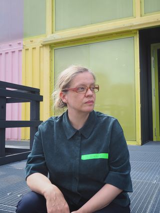 Portrait of Birgit Wudtke. She is seated in front of a yellow-green building with large window panels and looks to the right. She wears glasses and a dark shirt with a green name tag.