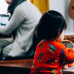 The photo shows a toddler at play and a mother at work on her laptop (both are pictured from behind).