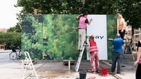 In »PostCompost – Forest Reset«, new large-scale posters are continuously layered onto a freestanding billboard in public space. Photo: Adam Schmidt Four people attach new image and paper panels to a large freestanding billboard in a public space. Two are standing on ladders, with trees and buildings visible in the background.