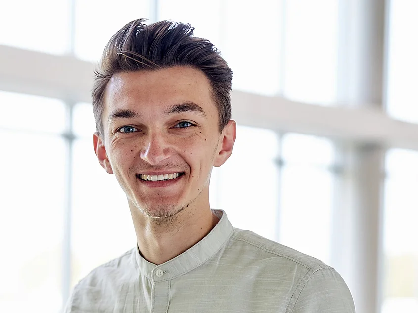 Portrait of Josha Helmchen. He smiles at the camera, wearing a light stand-collar shirt. Large windows in the background create a bright and open atmosphere.