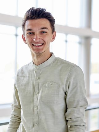 Portrait of Josha Helmchen. He smiles at the camera, wearing a light stand-collar shirt. Large windows in the background create a bright and open atmosphere.