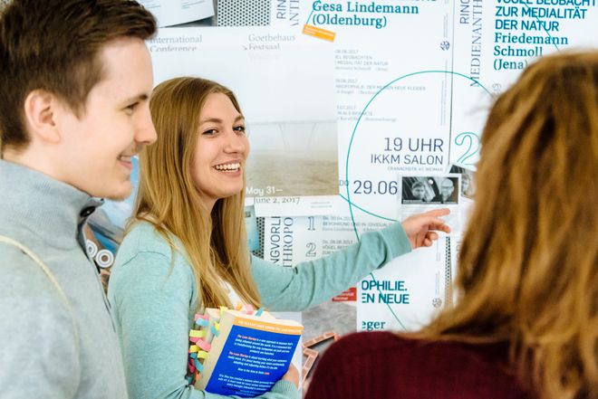 Das Bild zeigt zwei weibliche und einen männlichen Studierenden, eine der Frauen zeigt mit dem Finger auf eine Pinnwand mit universitären Plakaten (Copyright: Henry Sowinski)