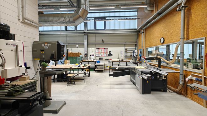 Großzügige Werkhalle mit stationären Holzbearbeitungsmaschinen, Arbeitstischen und Absauganlagen. Blick in einen technisch ausgestatteten Werkraum mit hohen Decken.