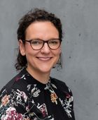 Porträtfoto von Tina Meinhardt vor einer grauen Betonwand: Sie hat mittellange, lockige braune Haare und trägt eine schwarze Bluse mit buntem Blumenmuster. sowie eine Brille mit dunklem Rahmen. Sie lächelt freundlich in die Kamera.