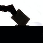 Das Foto zeigt eine Hand, die einen Wahlzettel in eine Wahlbox einwirft.
